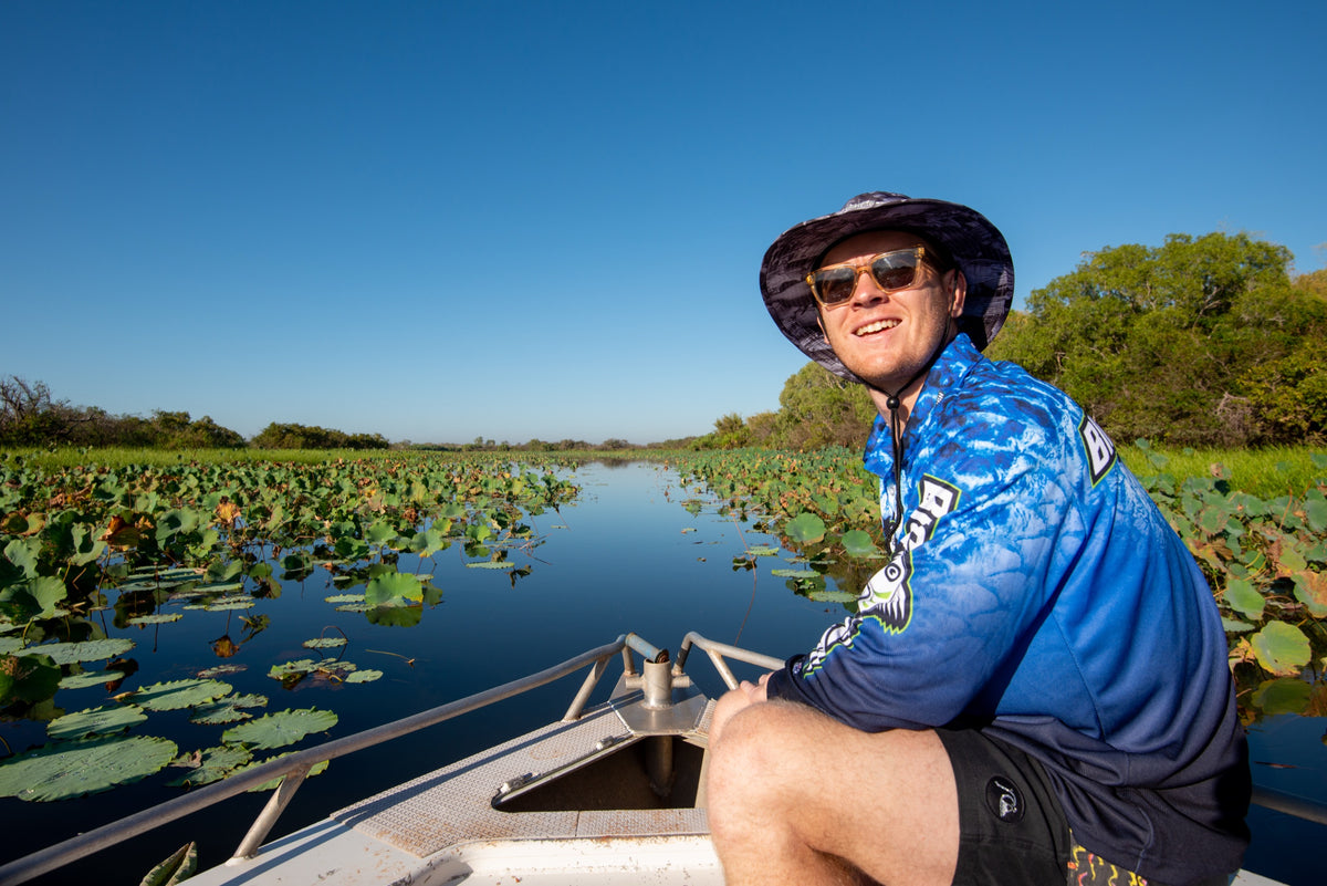 Angler in a Bigfish shirt and bucket hat on a lily-covered billabong